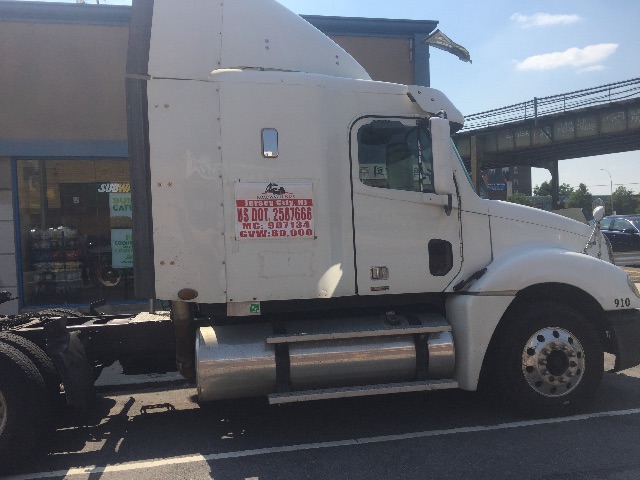 Truck side view showing white Freightliner Columbia sleeper cab with attached chrome fuel tanks. Visible stickers on door with small red text, and a number 910 on the front fender.
