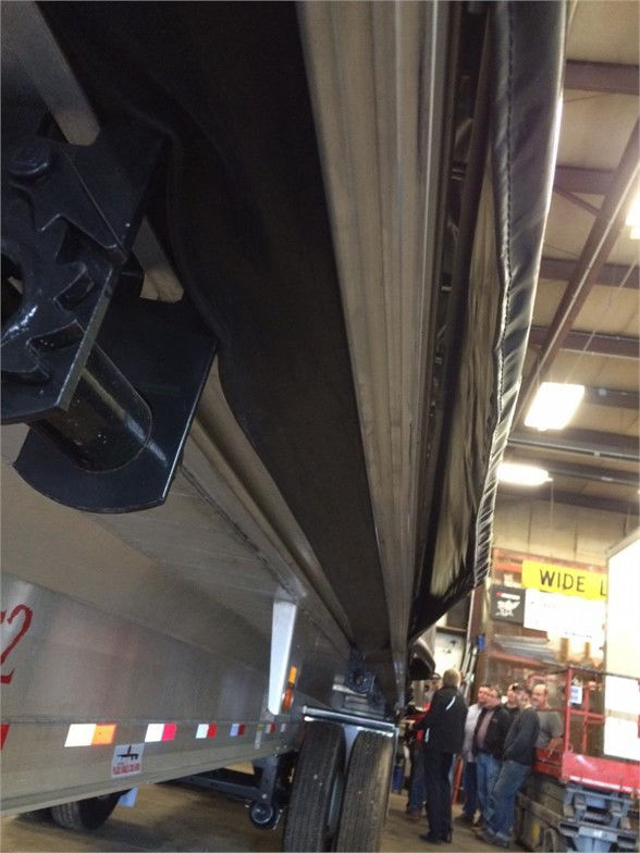 Trailer side view showing tarp system on Fontaine flatbed trailer with black tarp rolled along the rail and people in the background.