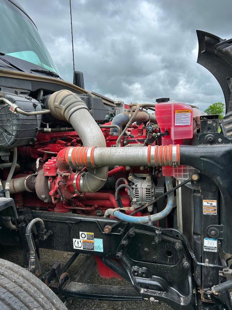 Kenworth T680 engine bay showing red engine components, large intercooler intake hose, coolant reservoir, alternator, and chassis with warning labels.