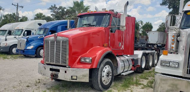 A red Kenworth conventional daycab tractor in a truck lot, front grille, large chrome bumper, dual exhaust stacks, and multiple trailers in background.