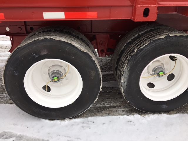 Semi trailer axles with white wheels and black tires, dual tandem configuration on a red trailer; visible snow and road grime around rims.