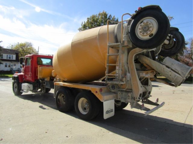 Concrete mixer truck rear view showing large tan drum and rear chute, tandem rear axles, and red cab in background.