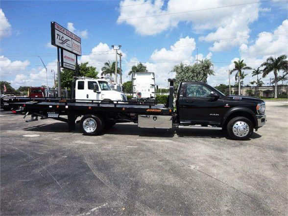 Ram 5500 Tow truck with a flatbed, black cab and chassis, large side mirrors, chrome wheels, stock number TBC, no visible decals readable.