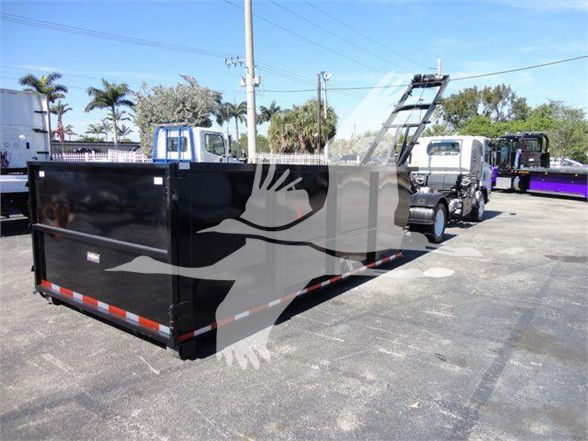 Isuzu NRR truck with a large black dump bed trailer attached, side view, in a lot with other trucks and palm trees.