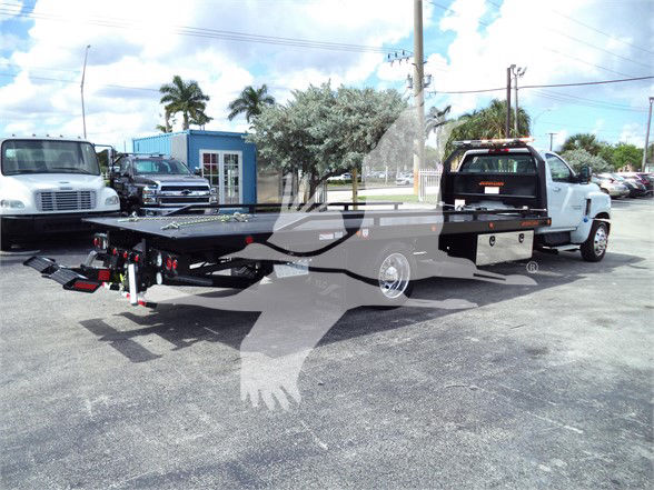 Flatbed tow truck trailer attached to white Chevrolet Silverado 6500HD with ramp and rear lights, in a dealership lot.