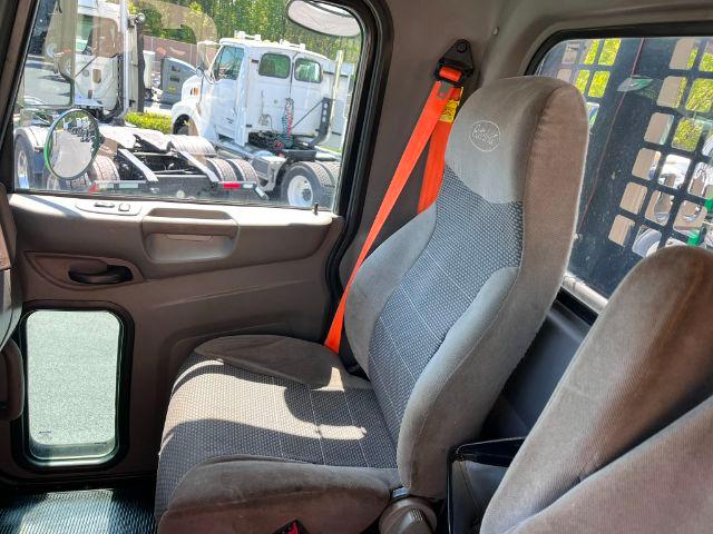Interior of a 2015 Peterbilt 348 cab showing driver seat with orange seatbelt, window, and partial view of exterior trucks.