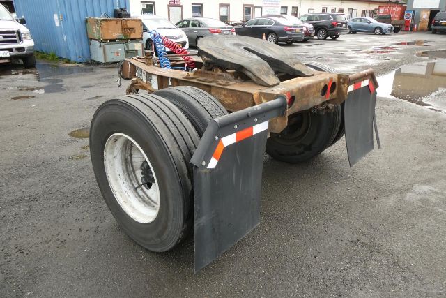 Trailer dolly chassis with dual tires, mud flaps, and visible steel frame; surrounding lot with vehicles and buildings.