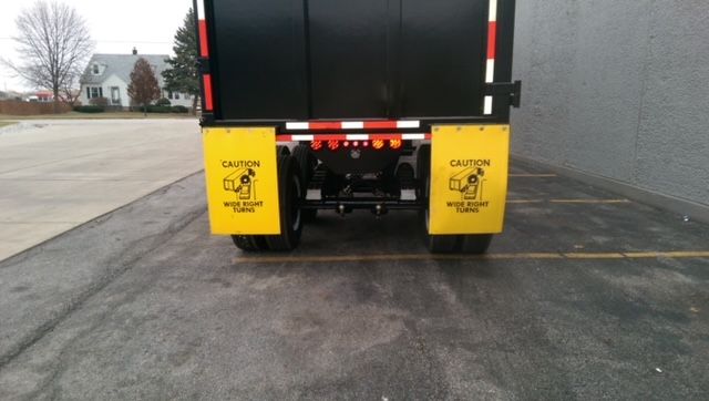 Rear view of a dump trailer with yellow safety mudflaps on each side, warning 'CAUTION' and 'WIDTH RIGHT' text, and red/white reflective stripes on the bumper. The trailer bed is black with wheels visible.