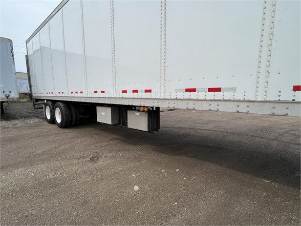 Trailer underside showing dual rear axles and undercarriage with storage box and landing gear, long white trailer with reflective red/white stripes.