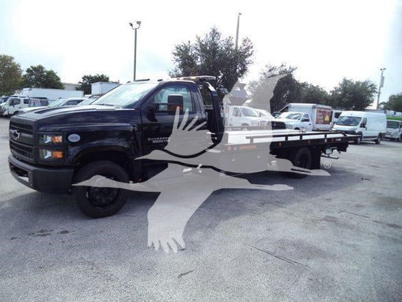 Truck with flatbed on a dealership lot, Chevrolet emblems visible on the black cab. Winged watermark across the image.
