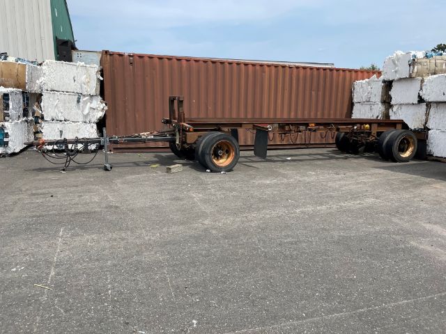 Open viewed trailer chassis with rusted frame and two axles, brown shipping container on frame, white blocks on either side.