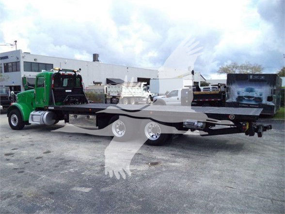 2014 Peterbilt 367 tow truck with a long flatbed trailer, green cab, black flatbed, twin rear axles, truckyard background.