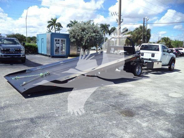 Flatbed tow truck with Ram 5500 chassis in a dealership lot, loading ramp extended, white truck in background.