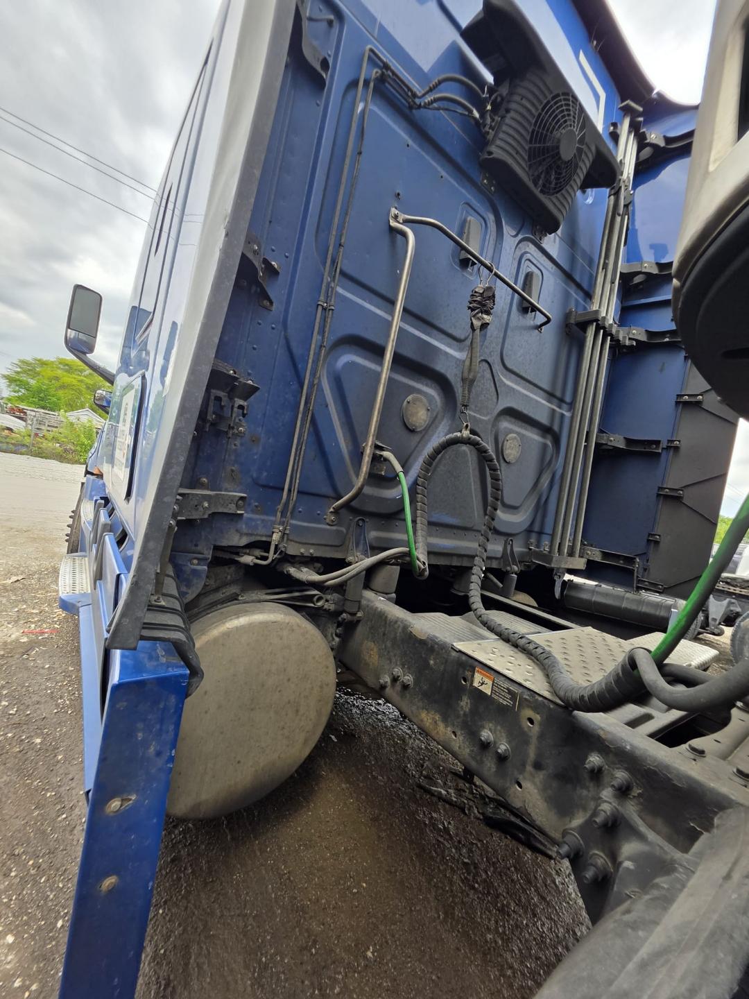 Truck cab rear wall of a blue Freightliner Cascadia with exposed wiring and hoses, circular fuel tank near ground, and chassis frame visible.