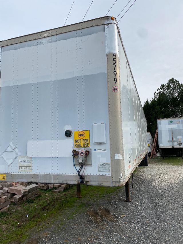 Trailer side view showing a white Wabash Duraplate trailer with vertical riveted panels, dual landing gear, and warning decals; appears to be parked in a lot.