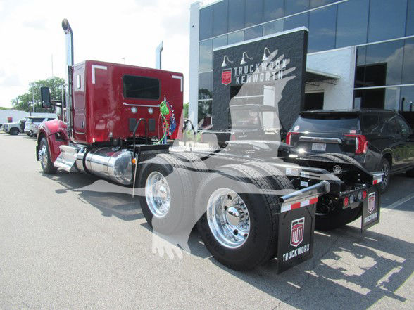 Kenworth W990 tractor truck in red sleeper cab seen from rear-left, showing dual rear axles, chrome wheels, tanks, and dealership building in background.