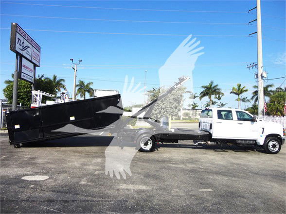 An armoured dump bed dump-truck attached to a white heavy-duty truck in a dealership lot; background shows palm trees, blue sky, and a signboard.