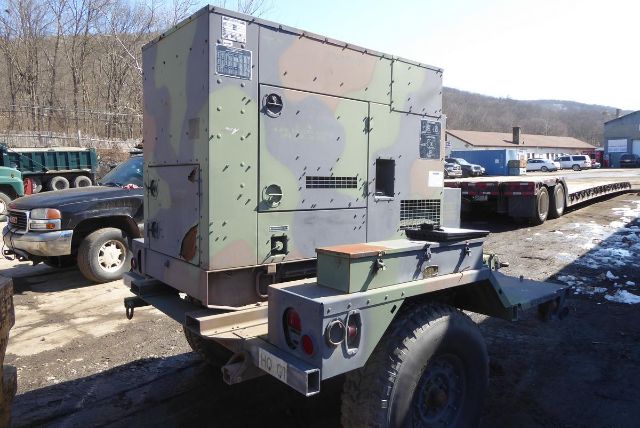 Camouflage-patterned military-style generator on trailer with wheels and hitch, mounted on a flatbed trailer in an outdoor yard.