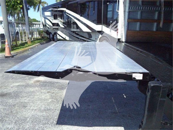 Flatbed trailer ramp extended from a Mack MD6 box truck, loading area visible with metallic surface and black hinge mechanism at edge.