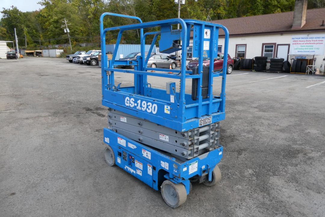 GS-1930 scissor lift in blue with stacked sections and control panel, side label GS-1930, wheels on asphalt, dealership backdrop.
