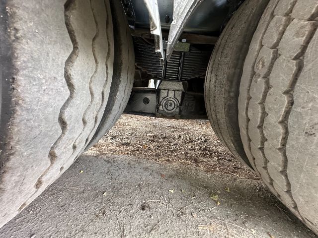 Underneath a truck showing the hitch area between dual rear wheels. Visible components include a center crossmember, suspension hanger, and a small circular cap with possible bolt patterns.