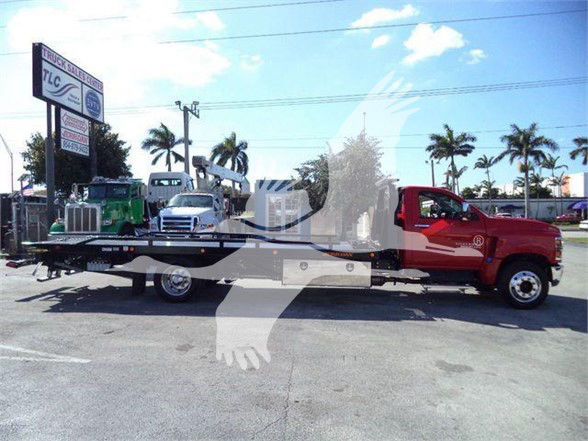 Truck mounted tow vehicle with red cab and flatbed, Chevrolet Silverado 6500HD, on dealership lot with palm trees.