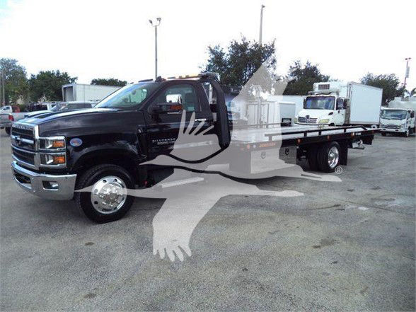 Tow truck with flatbed in black Chevrolet Silverado 6500HD, front grille, dual rear axles, dealership lot in background.