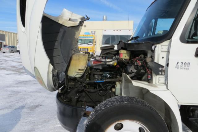 Box truck engine bay of a 2003 International 4300 showing exposed engine components and front wheel. Visible labels include '4300' on the door and various hoses and reservoirs.