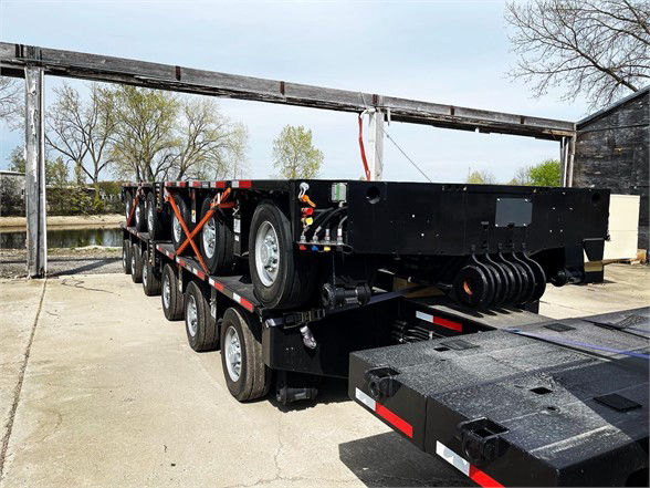 P-12 trailer side view showing heavy-duty double drop deck trailer with multiple axles, spare tire, and hydraulic ramps. Visible components: black steel deck, wheel groups, reflective tape, and a coiled mechanism at the rear.