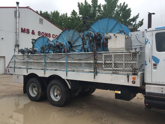 Truck bed with large reel-like spools on a service/utilities truck, dual rear wheels, aluminum side railing, white cab, industrial area background