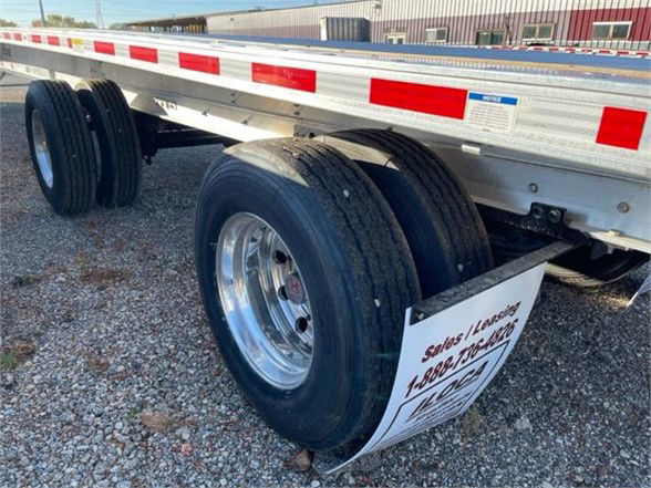 A Fontaine flatbed trailer showing dual rear axles with chrome wheels and black tires; white reflective side rails; a white sign hanging near the wheel with handwritten text.