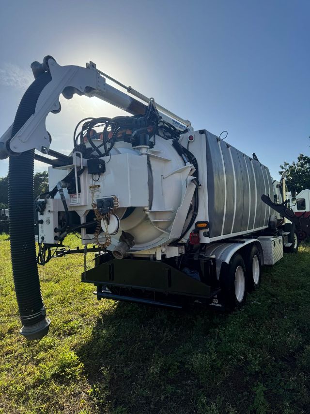 Truck-mounted vacuum tanker with large rear fill hose, white tank, dual rear axles, and hydraulic arm. Side view shows metal panels and electrical wiring.