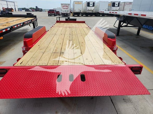 Flatbed trailer with wooden deck and red steel ramp sections at the rear, parking lot with other trailers in background.