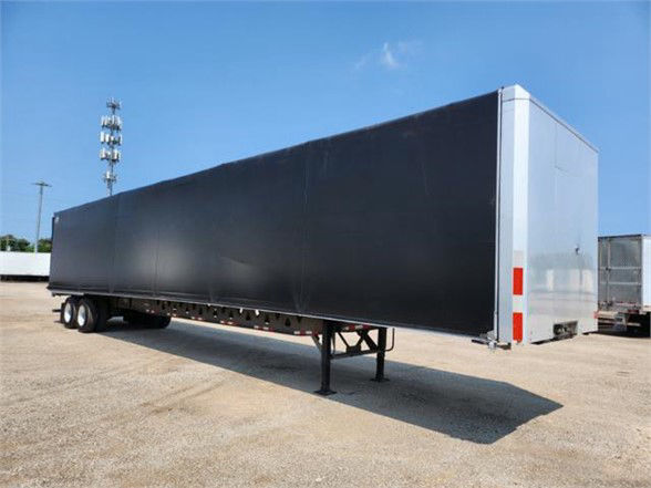 Black Manac combo flatbed trailer viewed from side with gooseneck and dual rear axles, standing on a gravel lot.