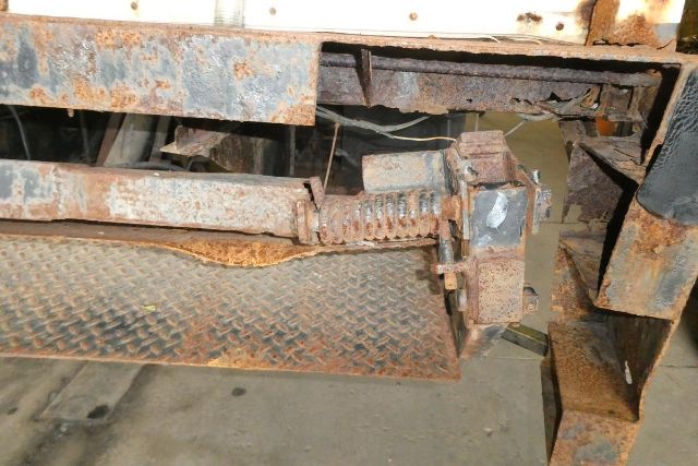 Box truck undercarriage with rust and mechanical linkage, showing a rusty folding step and linkage near a diamond-plate platform in a Freightliner M2 106 chassis.