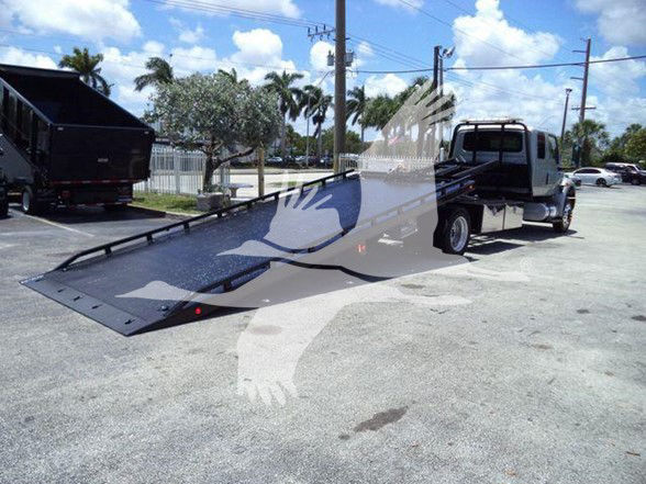 Tow truck with a long black tilt bed trailer in an outdoor lot, International MV rear view showing ramp bed extended.