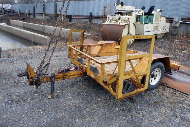 Cross Country trailer with yellow metal frame and hitch, attached chains, and a detached drum roller in the background; rugged outdoor lot.