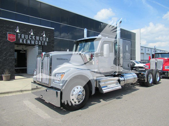 Kenworth W990 conventional sleeper semi-truck in showroom lot, chrome exhaust stacks, large grille, front bumper, and cargo area visible.