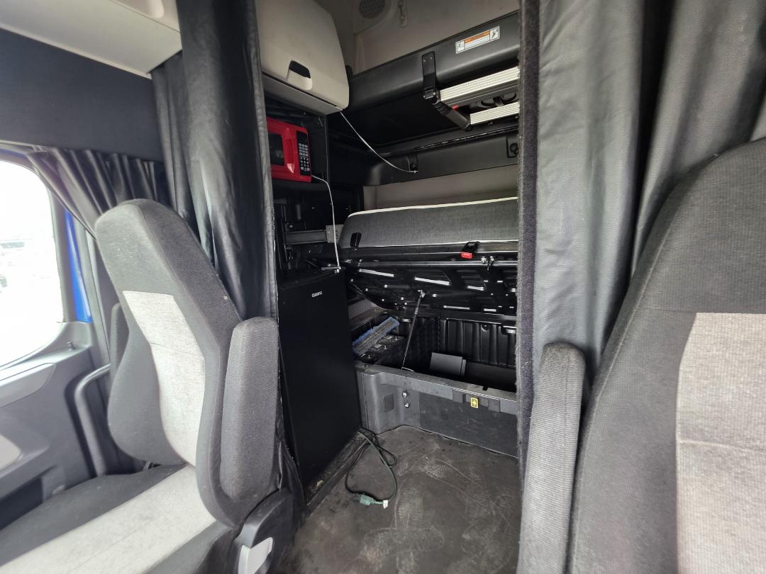 Cab interior of a 2018 Freightliner Cascadia tractor showing a sleeper bunk with a small refrigerator, wiring, and storage compartments visible behind the driver's seat.