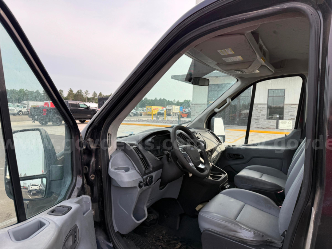 Ford Transit commercial van interior front cabin with steering wheel and driver seat, gray dashboard, and passenger seat visible; side mirrors and windshield show exterior lot in background.