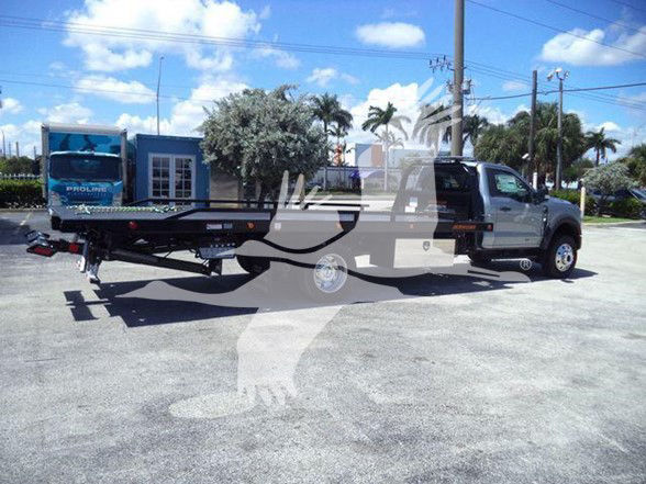 Ford F-600 tow truck with a flatbed trailer attached to a gray/black cab. Visible text includes dealership branding on the trailer and a pickup bed area; no legible model-specific stickers.