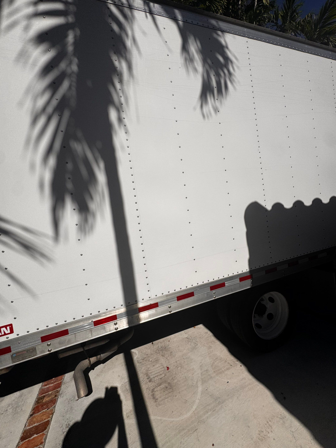 Trailer side panel with riveted aluminum walls and a wheel in shadow. Red/white reflective tape along lower edge; exhaust pipe/curved tube near bottom left.