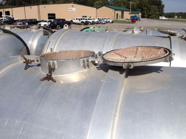 Industrial vacuum tank truck top view with circular access hatches and clamped rings, metal surface reflecting sunlight; background shows a service yard with buildings and parked trucks.