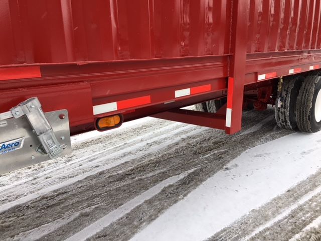 Side of a red trailer with white and orange reflective strips, undercarriage and lights visible. Snow on ground.