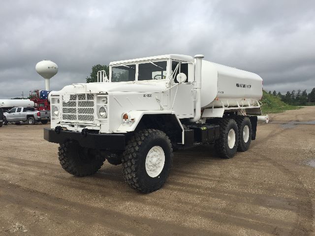 M923A1 military water tanker truck showing large white tanker with dual rear axles and heavy off-road tires. Front grille, round headlights, and raised cab. No visible text on side of tank.