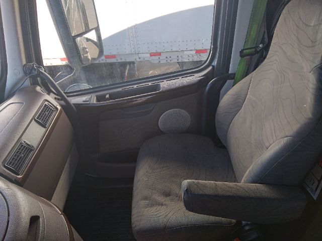 Truck cab interior with steering wheel on left, dashboard vents, and a fabric seat. Visible through window is a trailer.