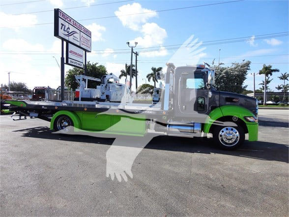 Tow truck with green and silver paint, long flatbed, Peterbilt emblem on front. Visible signage in background, large white silhouette watermark over the image.