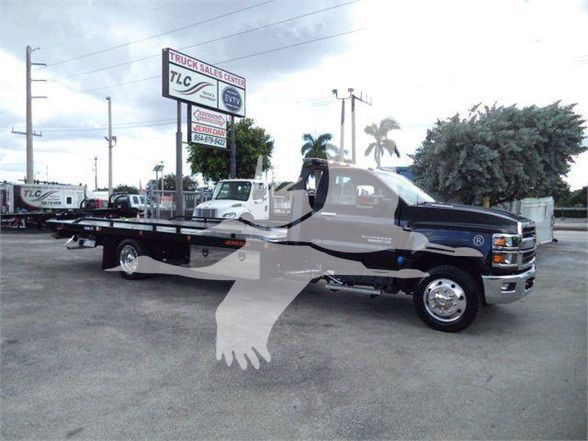 Truck bed tow truck chassis of a 2023 Chevrolet Silverado 6500HD with extended flatbed and dual rear wheels, dealership lot in background.