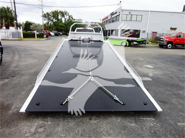 Flatbed tow truck bed on a 2023 Ram 5500 HD SLT with ramp extended, chains securing cargo, dealership lot in background.