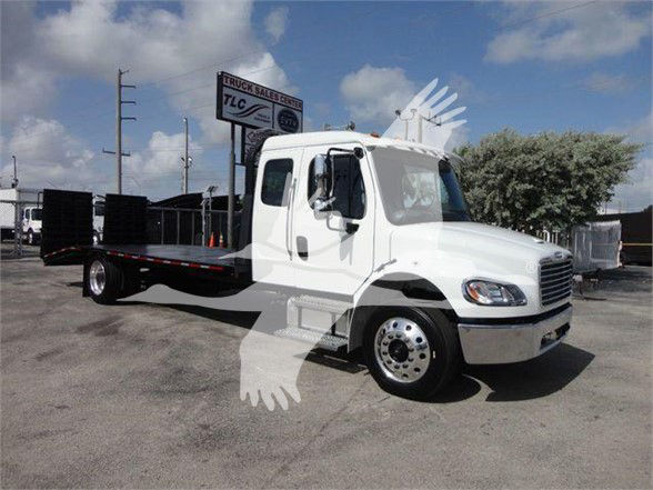 Flatbed tow truck in white with cab and long black flatbed. Visible text on sign in background: TLC Truck Sales & Service. The image shows a Freightliner M2-106 cab with chrome wheels.